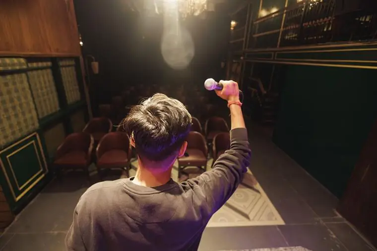 A young performer seen from behind on a small theatre stage, microphone raised toward the empty seats: reading the room