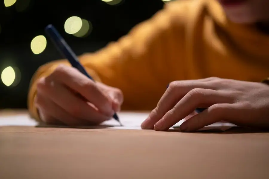 A hand writing on a sheet of paper, warm lights blurred behind: keeping a joke notebook