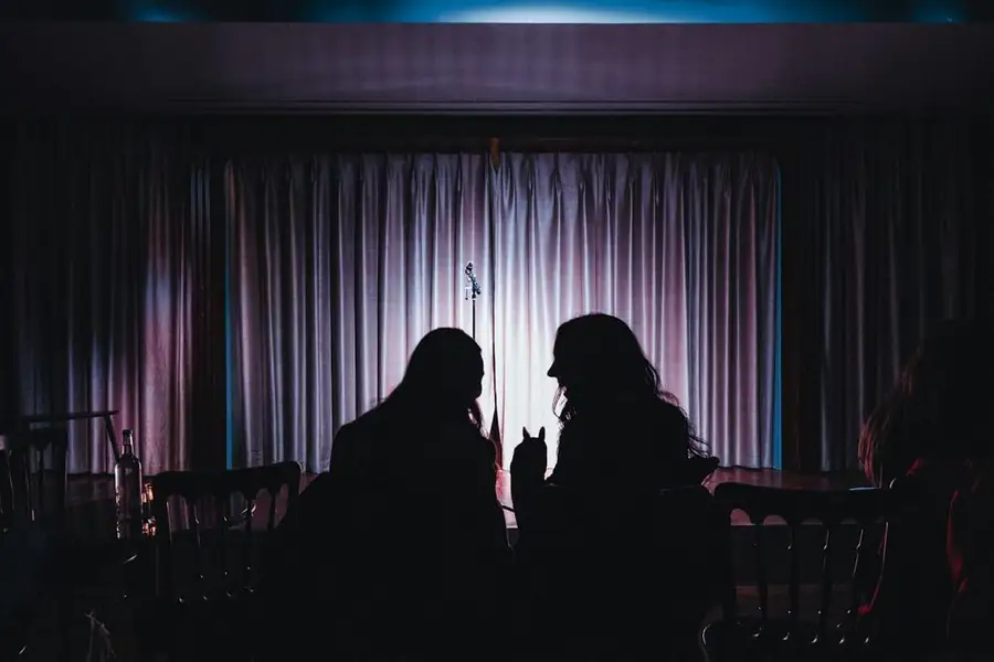 Two people in silhouette talking at a table in front of a lit stage curtain and mic stand: what a comedy agent does