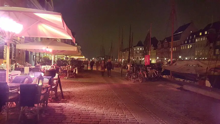 Stand-up comedy in Denmark: Nyhavn in Copenhagen at night, café umbrellas lit pink along the cobbled quay and old ships moored by the lit houses