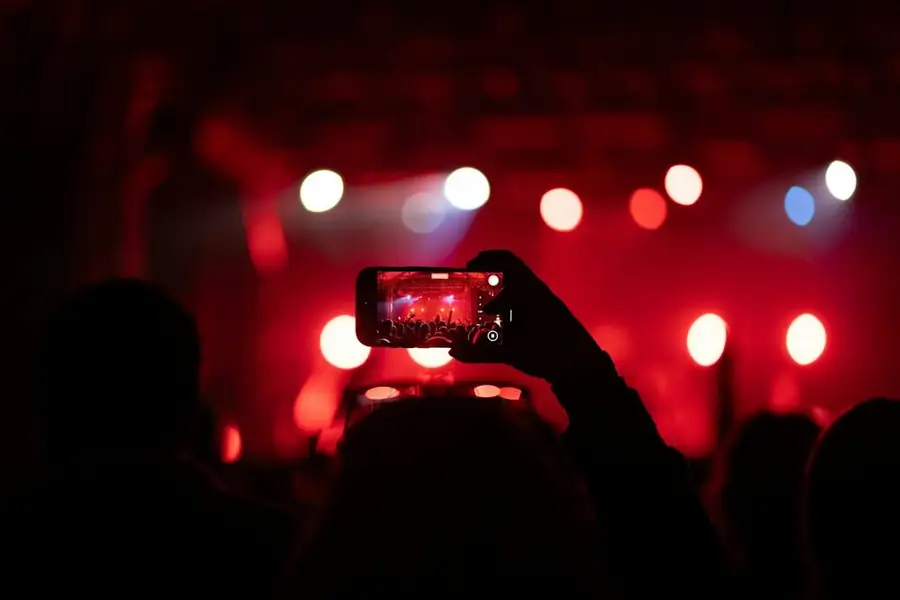 A phone held up in the crowd films a comedy stage in red light, for a comic about to submit a stand-up comedy clip to the Comedytron