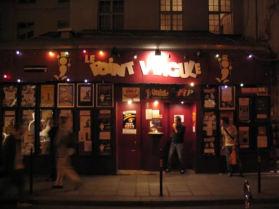 Stand-up comedy in France: the lit front of Le Point Virgule in the Marais, Paris, at night, its name in white above a doorway hung with posters and coloured lights