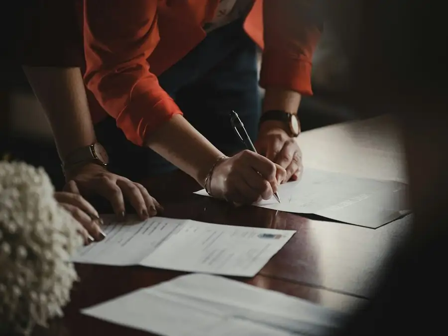 Two people leaning over a table, signing papers together; hands and forearms only: comedy contracts