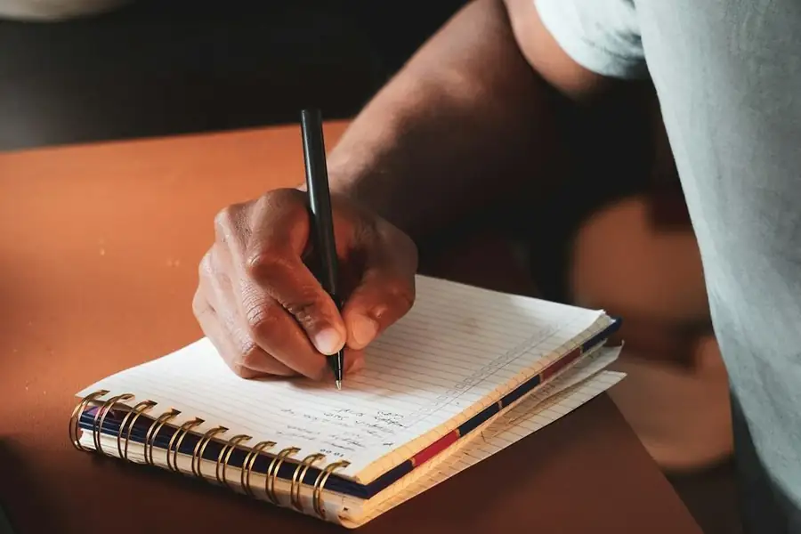 A hand writing in a spiral planner on a warm orange desk: writing a comedian bio