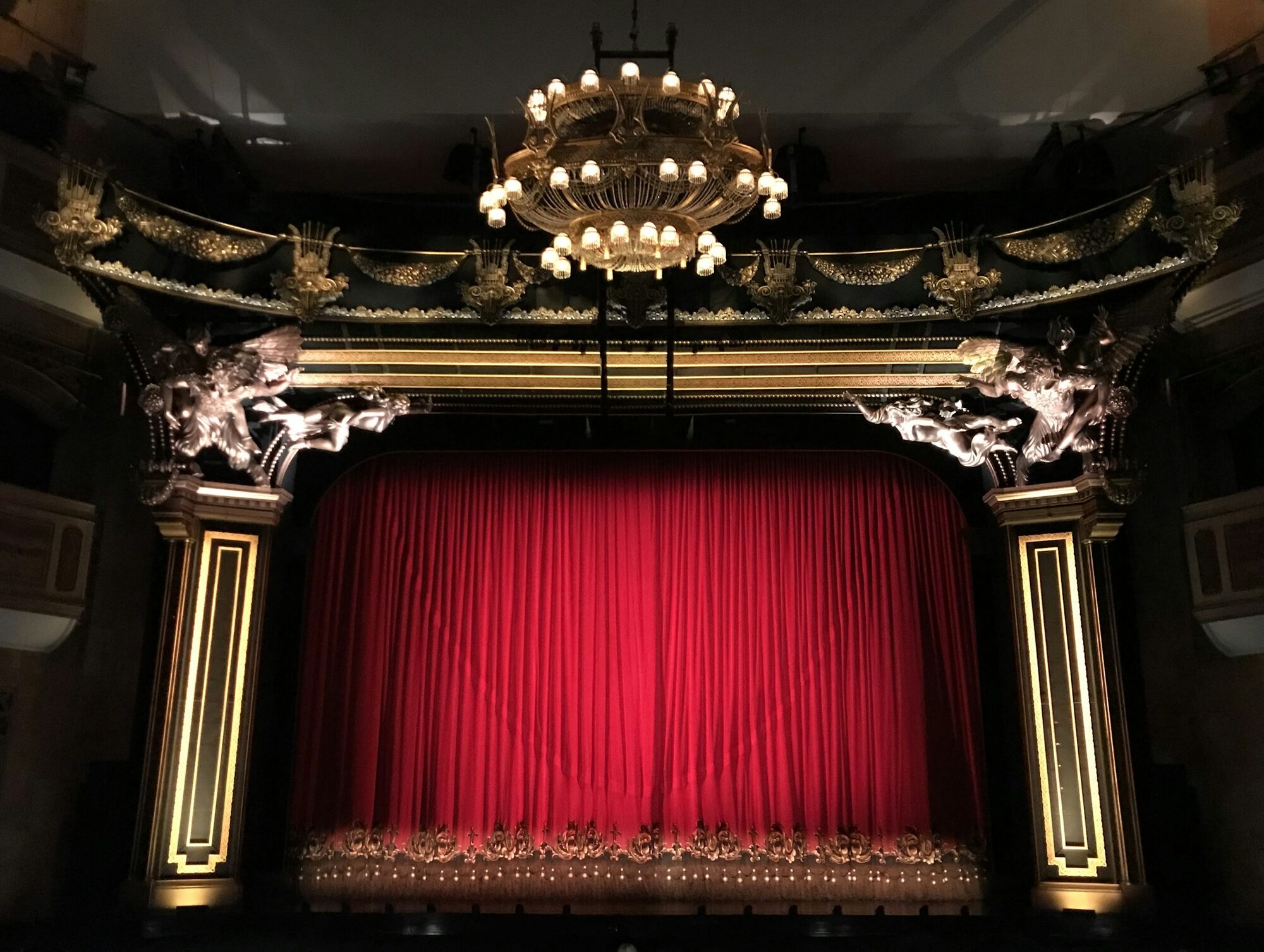 A stage fit for the greatest comedians: an old theatre's ornate proscenium, gilded carved figures, a chandelier and a closed red curtain