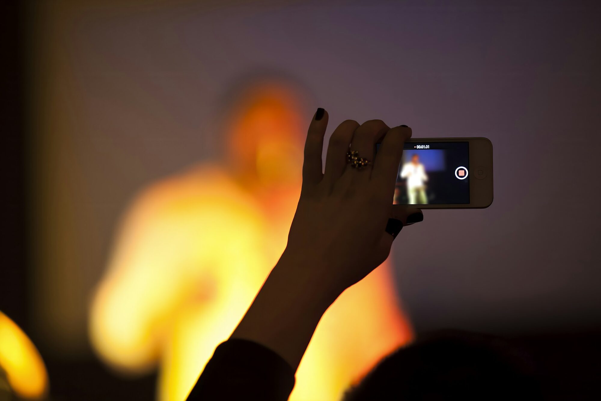 A hand holds up a phone to film a show, the stage a warm blur behind: one way to promote your comedy show