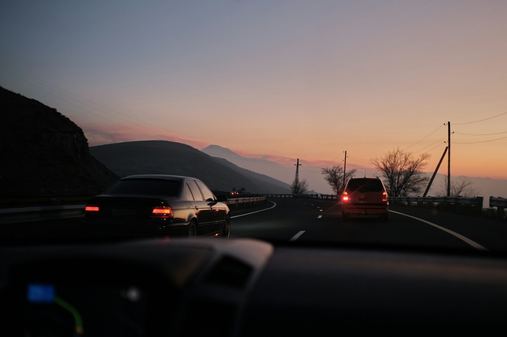 The road a comedy tour planner maps: dusk from the driver's seat, a car ahead with its tail-lights on, hills and power lines against a pink-orange sky