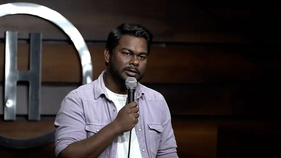 Stand-up comedy in India: comedian Haseeb Khan at the microphone on the stage of The Habitat in Mumbai, its wood-panelled wall and silver H behind him