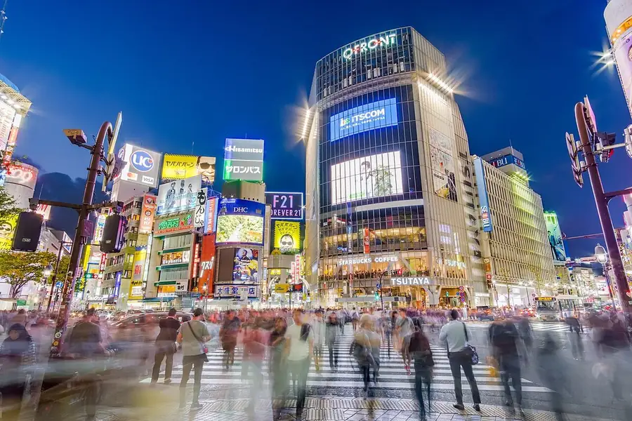 Stand-up comedy in Japan: the Shibuya Scramble Crossing in Tokyo at dusk, crowds crossing under lit screens and signs, by Shibuya Station, home of Tokyo Comedy Bar