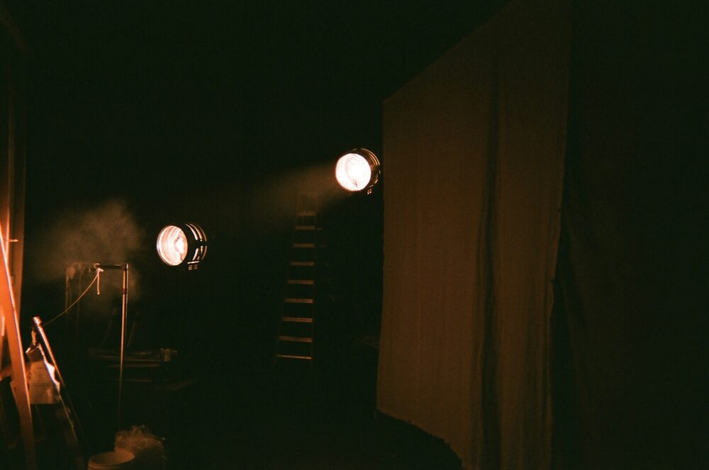 Backstage in the dark, where stage fright starts: two old spotlights glowing through haze beside a curtain, a ladder and a stand in shadow