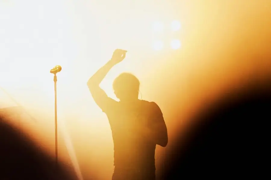 A performer in silhouette on a big stage, arm raised, a mic stand beside him, flooded with amber light: what a comedy headliner does