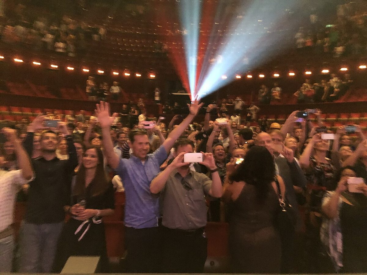 Stand-up comedy in the Netherlands: a cheering audience on its feet in Amsterdam's Carré theatre after Boom Chicago's 25th-anniversary reunion show, stage lights sweeping the hall