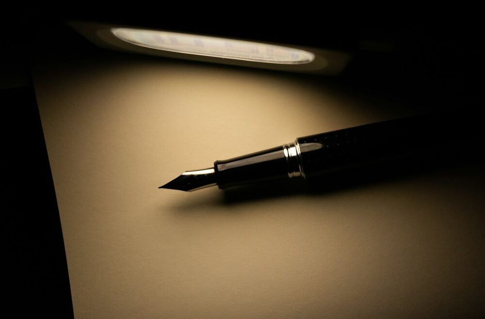 A blank page for learning how to write stand-up comedy: a fountain pen on a sheet under a desk lamp, everything else in shadow