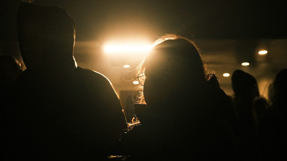 Comedy club etiquette from the audience's side: two people seen from behind, silhouetted against warm stage light
