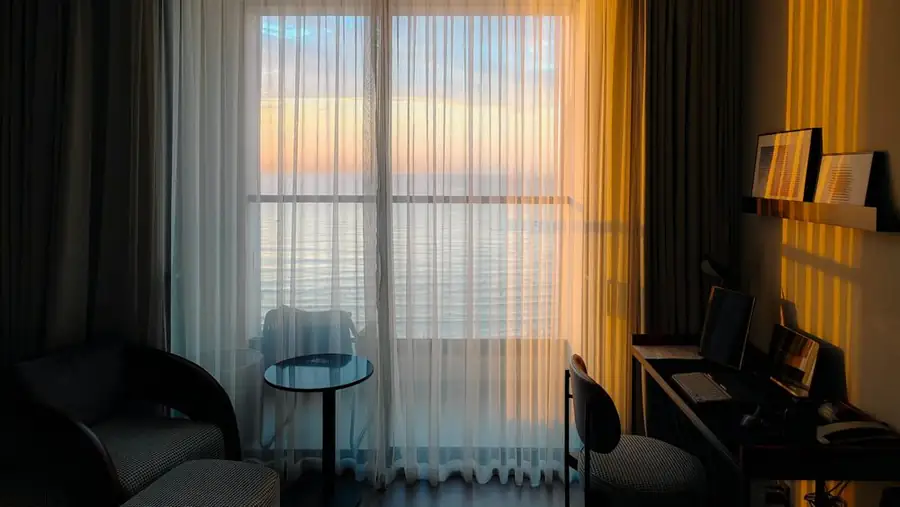 A hotel room in late-afternoon light: sheer curtains over a sea view, an armchair and a desk: the comedian's packing list