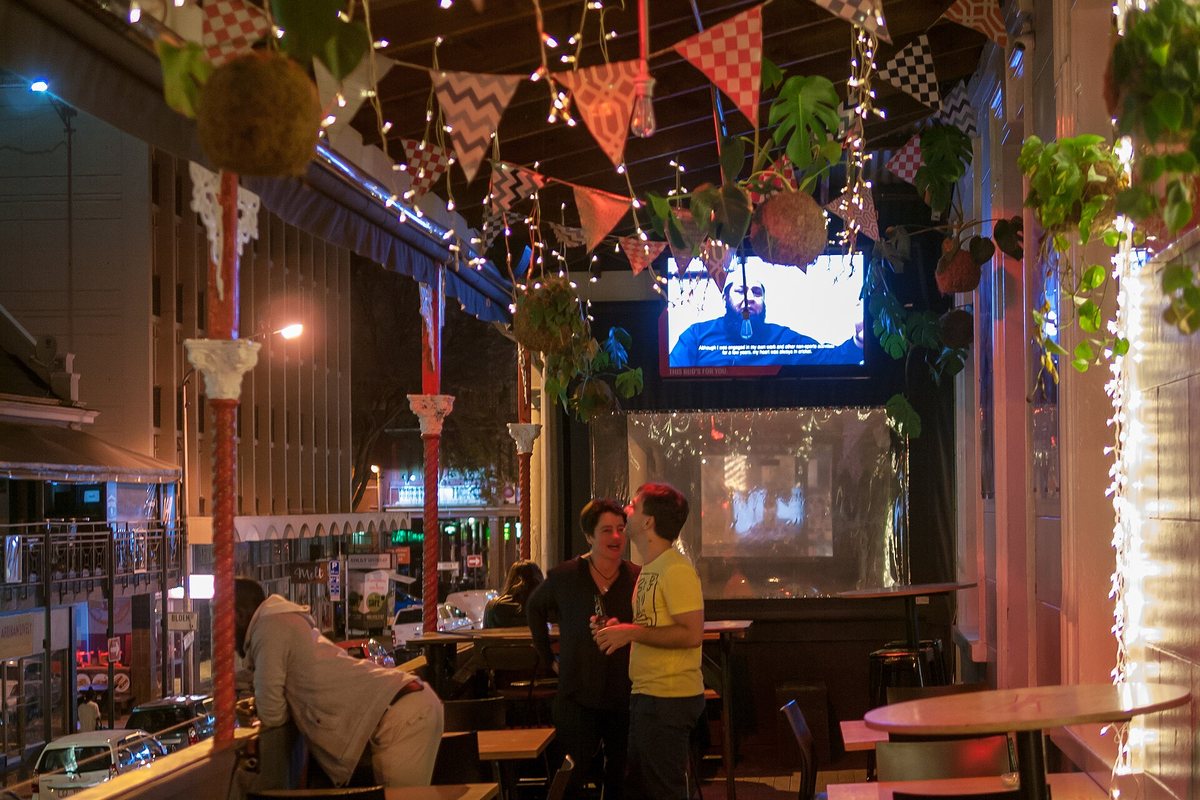 Stand-up comedy in South Africa: a Cape Town bar's balcony at night, strung with fairy lights and bunting, friends talking between the tables